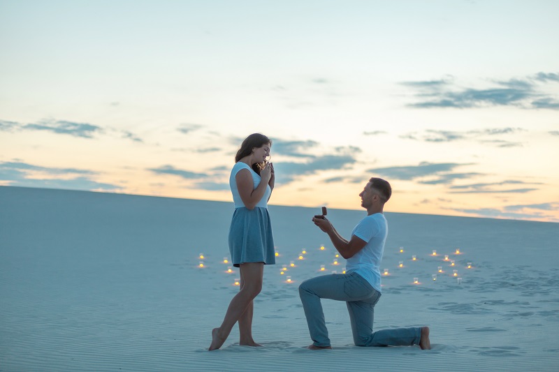 The guy makes the girl a marriage proposal by bending his knee while standing on the sand in the desert. Evening, candles burn in the sand.