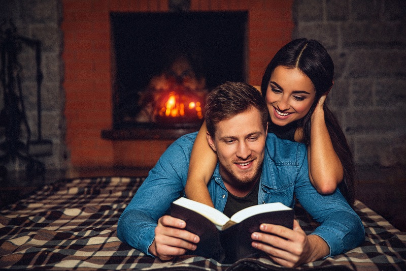 Positive young man lying on floor and read opened book. Beautiful model is behind him. She looks down at book and smile. They spend time together near fireplace
